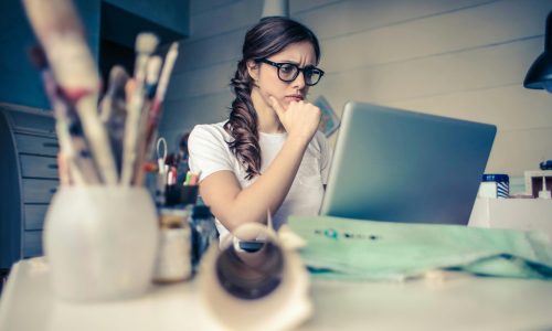 Young woman with glasses deeply focused on a laptop surrounded by art supplies in a home office.
